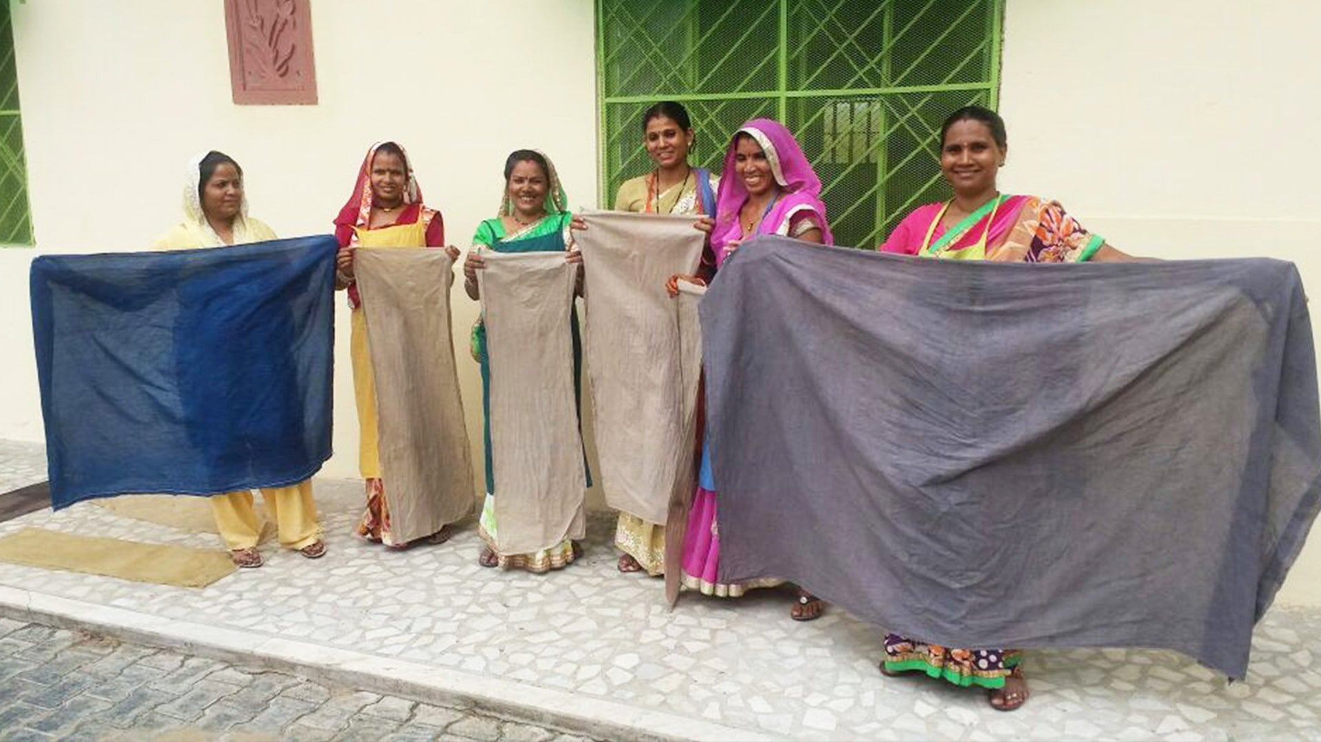 Six Anchal artisan women each holding up naturally dyed fabric from the project Designing colorful change, provided by the grant from Dining for Women