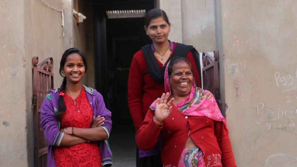three Indian women outside of their home waving at the camera