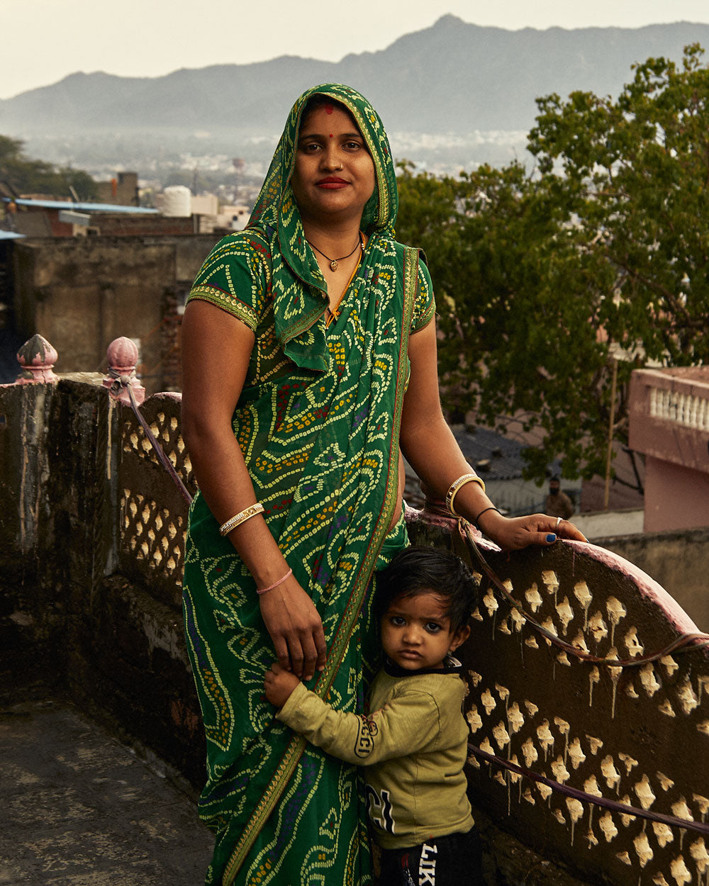 Woman in a green saree holding a child on a rooftop with mountains in the background