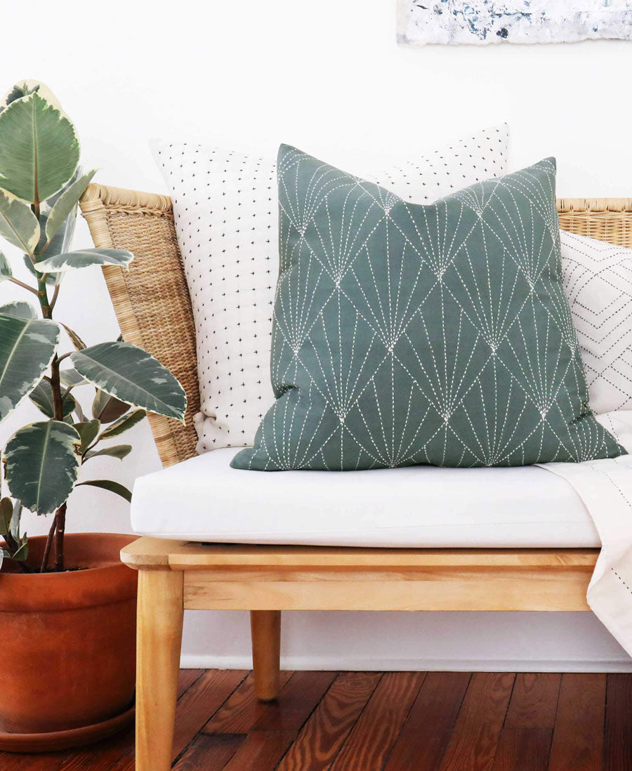 spruce green array throw pillow sitting on white and wicker entryway bench with plants in the background