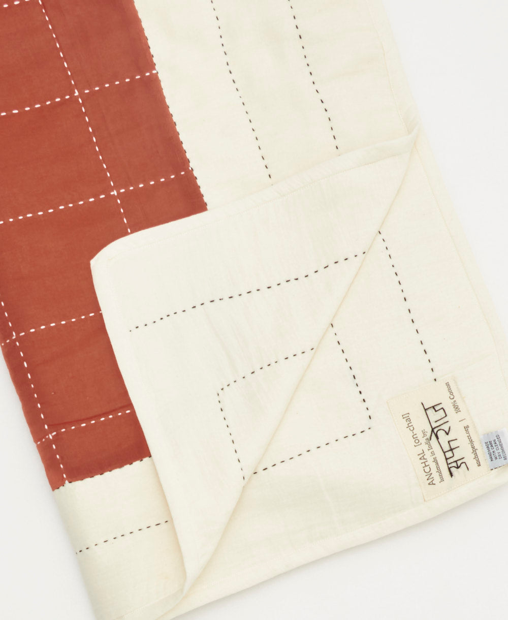 Close-up of a quilt with rust red and off-white panels on a white background, featuring the brand Anchal