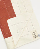 Close-up of a quilt with rust red and off-white panels on a white background, featuring the brand Anchal