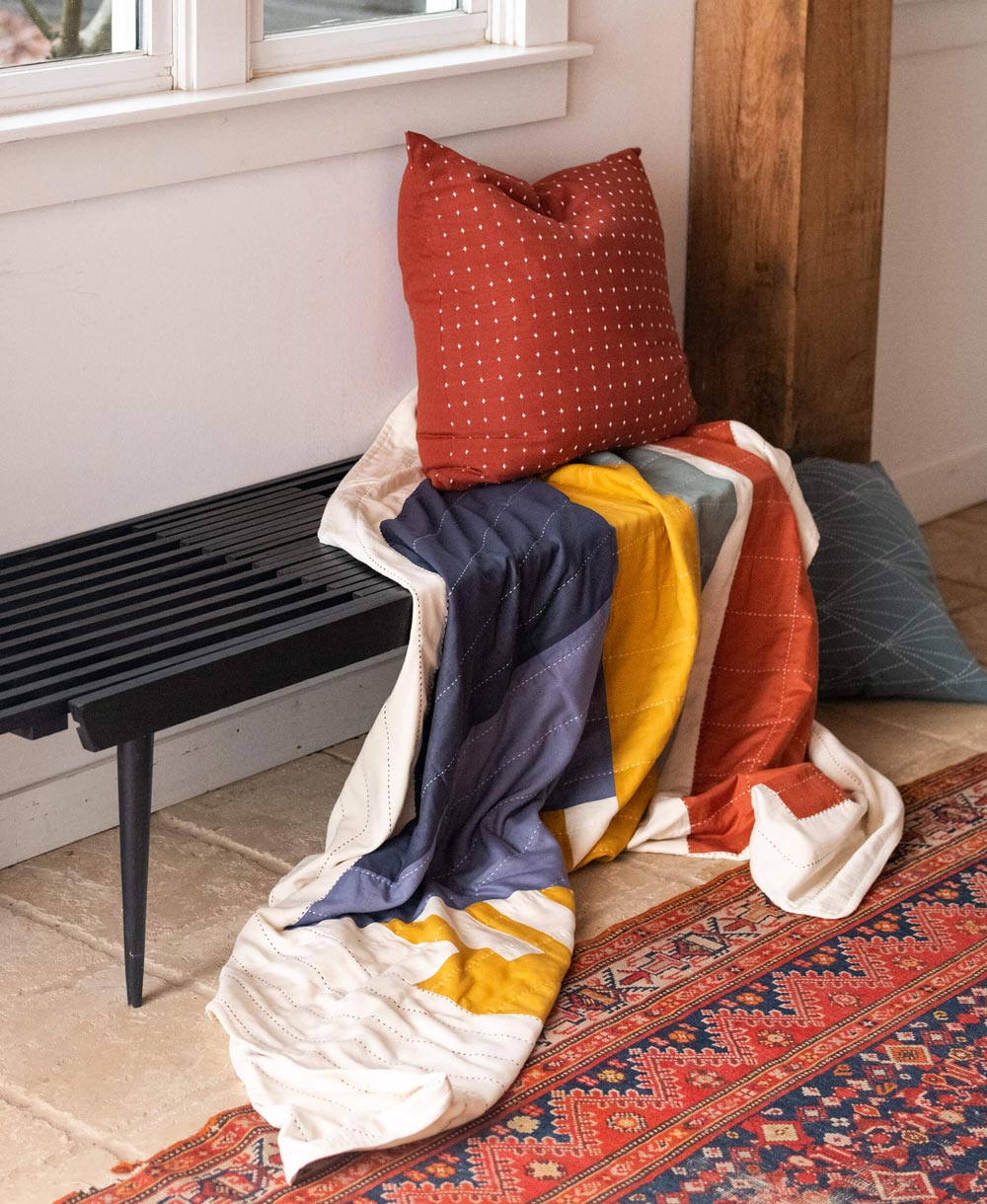 Colorful quilt draped over a black bench with pillows on a patterned rug.