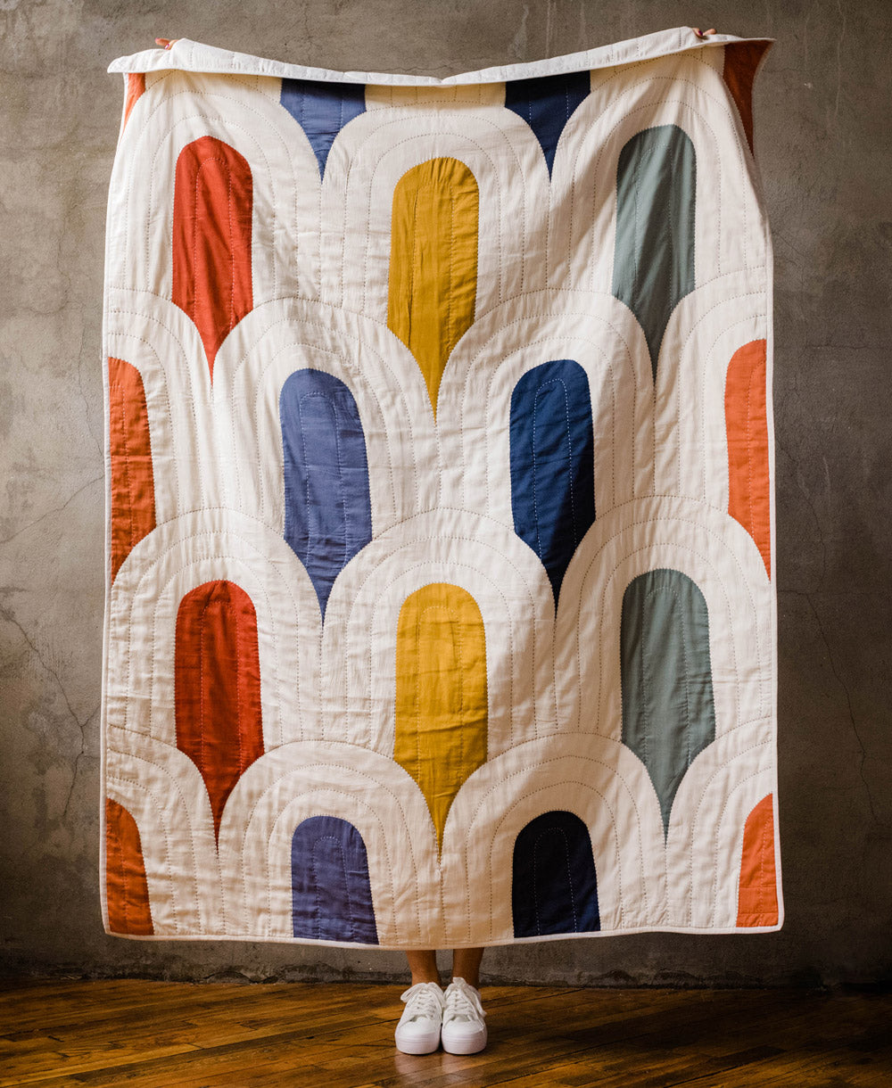 A colorful quilt with a geometric pattern is draped over a person against a textured wall