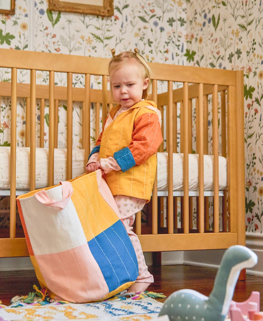 Child holding a colorful patchwork fabric storage bin in a nursery with floral wallpaper.