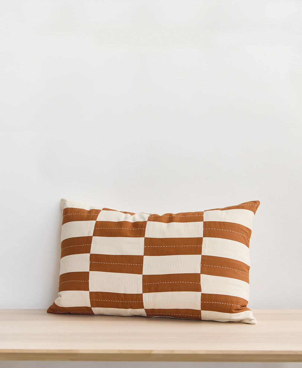 Checkered lumbar pillow with camel brown and white stripes on a light wooden surface against a white background