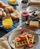 Breakfast table with waffles, fruit, and a jar of jam on a patterned tablecloth