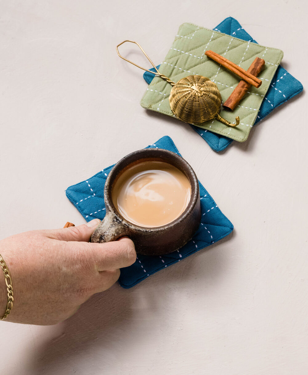 Hand holding a mug of coffee on a blue quilted coaster with a green coaster featuring a pumpkin latte and cinnamon sticks