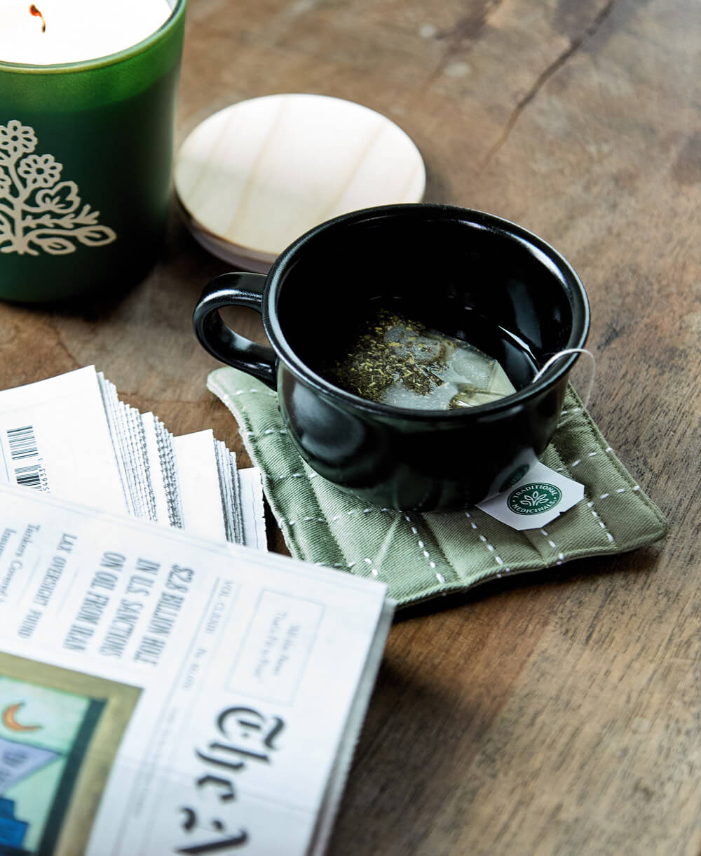 Black mug with tea bag on a green coaster next to a green mug and newspaper on a wooden surface