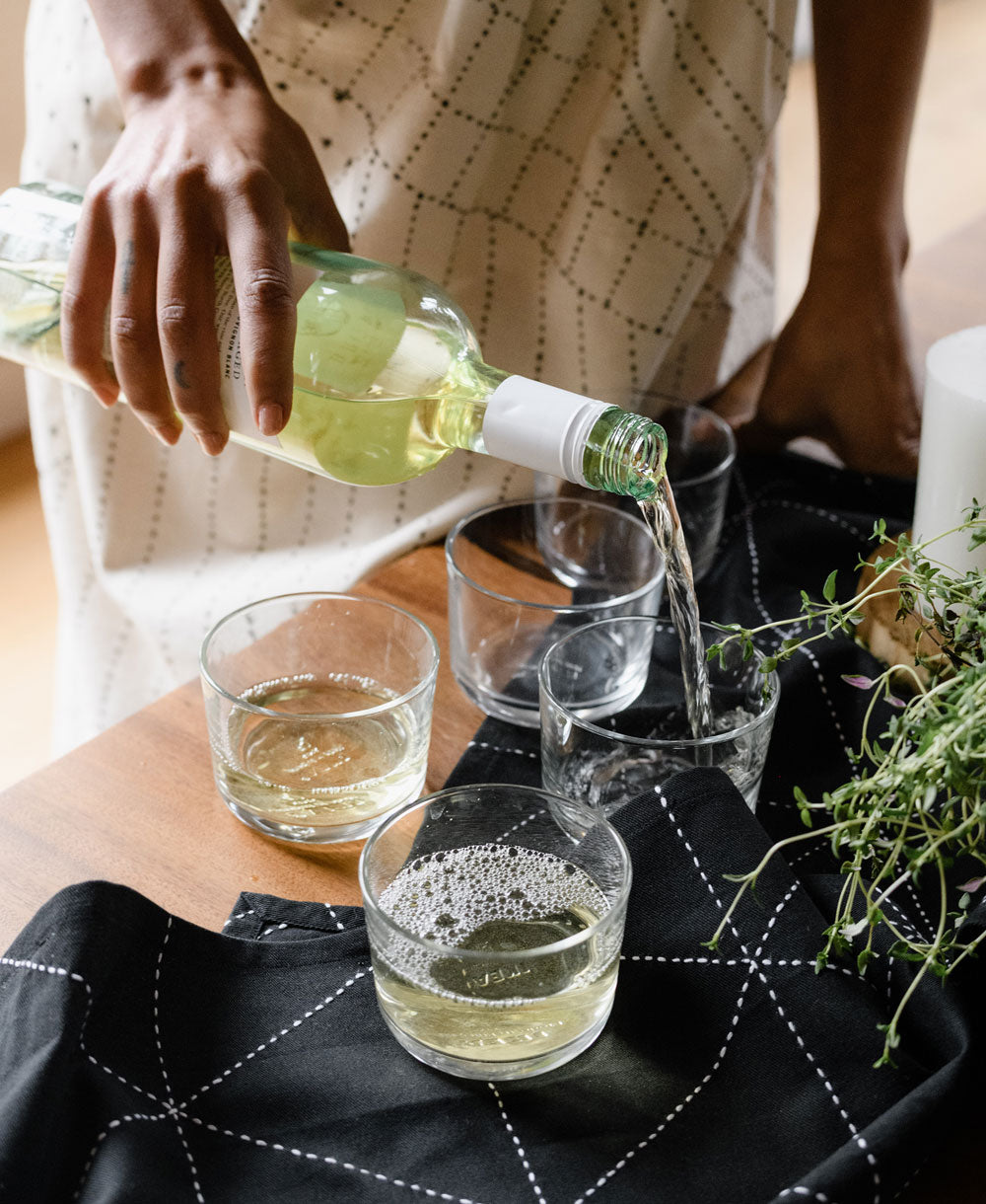 Person pouring white wine into glasses on a table with a candle and plant