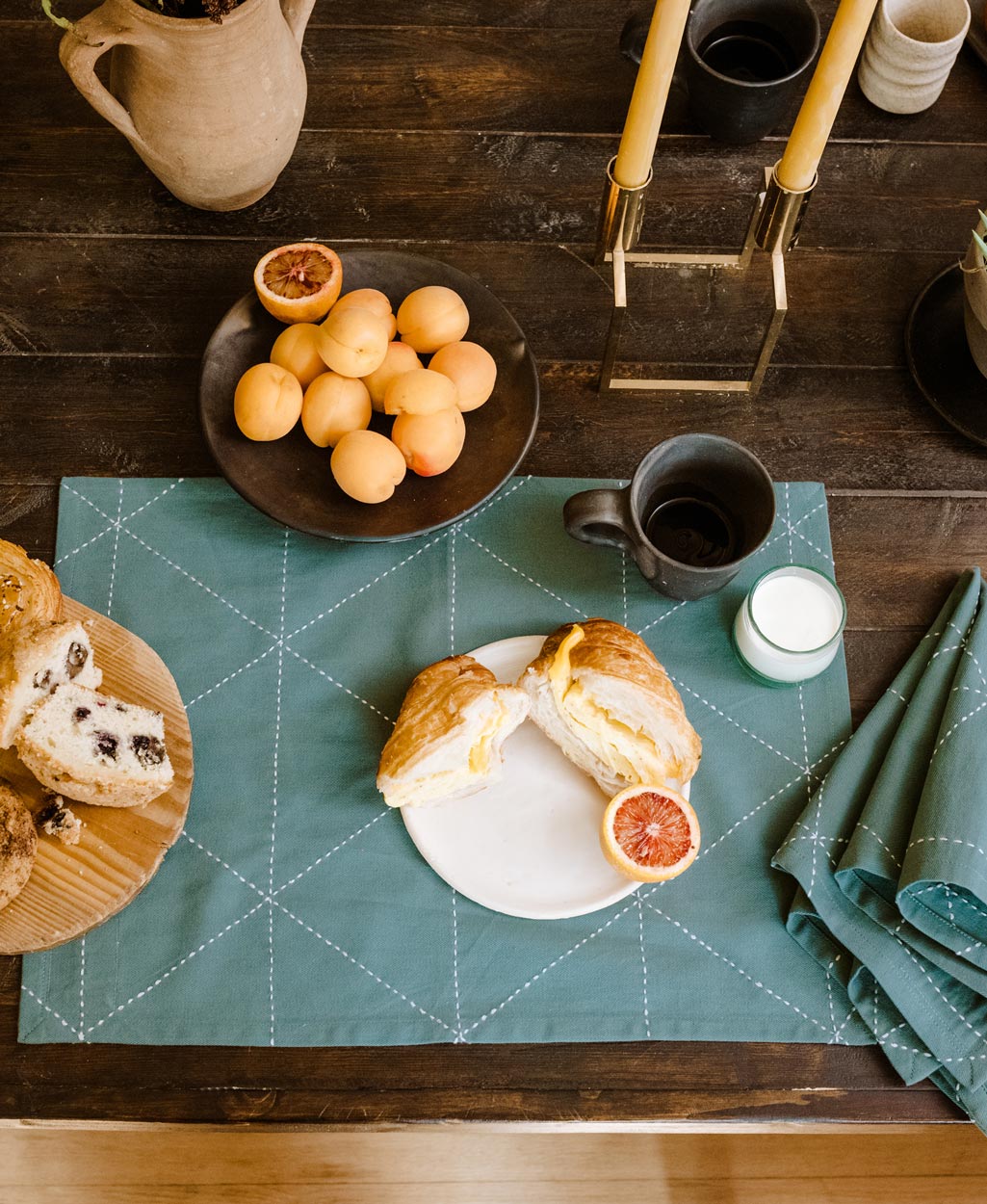 Breakfast setting with croissant, oranges, and coffee on a rustic wooden table with green cotton placemat