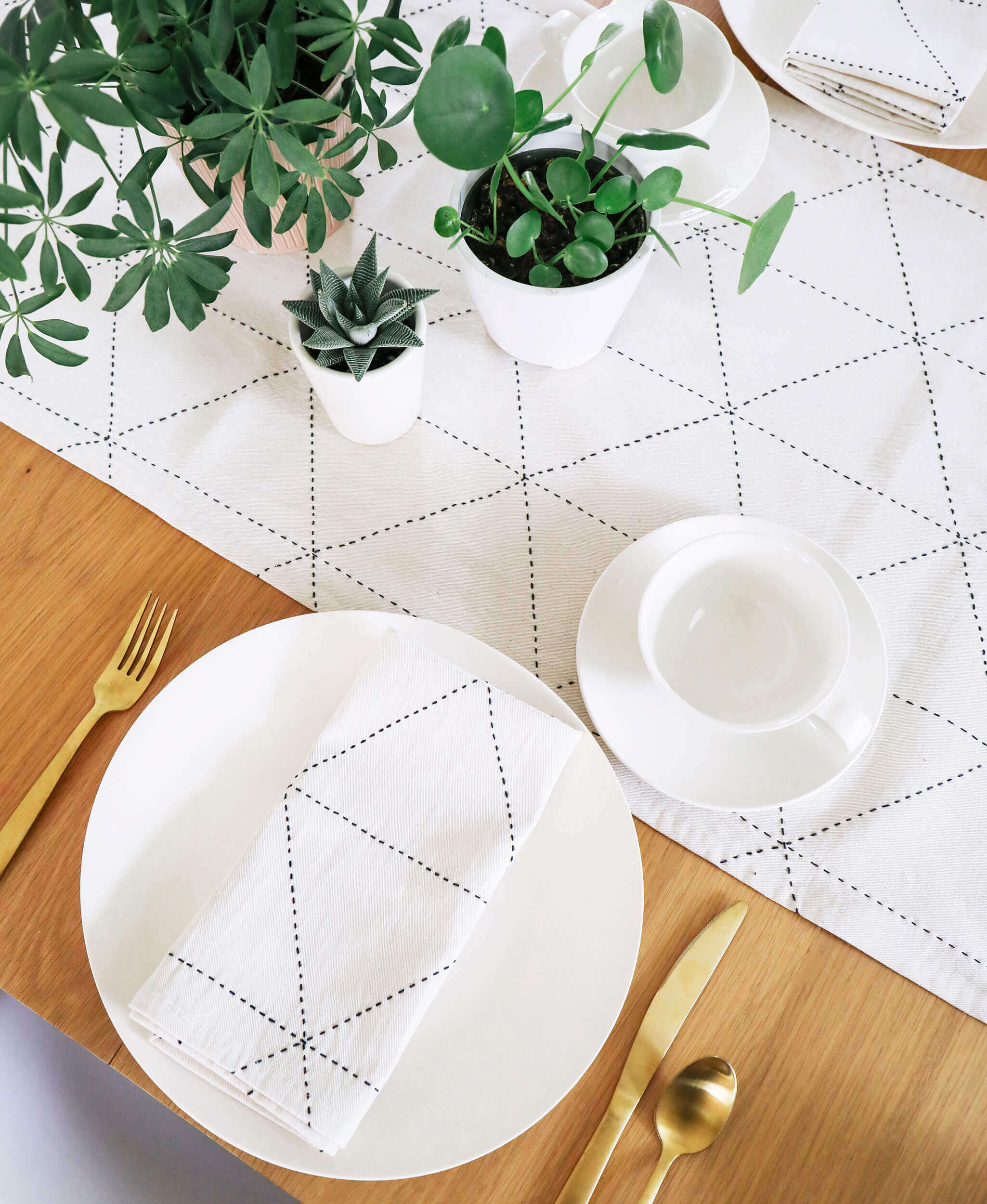Dining table setting with white plates, gold cutlery, and potted plants on a wooden surface