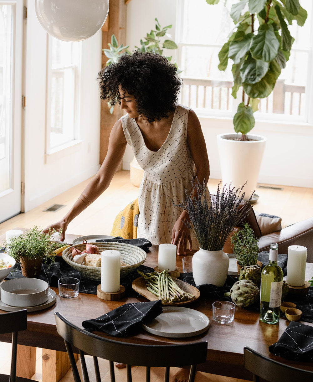Woman setting a table with black table linens with a decorative arrangement of plants and candles in a bright room