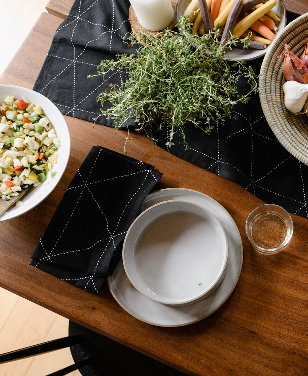 Dining table setup with plates, a bowl of salad, and a basket of vegetables on a black and white embroidered tablecloth