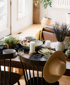 Dining table set with black table linens, candles, plants, and a hat in a bright room