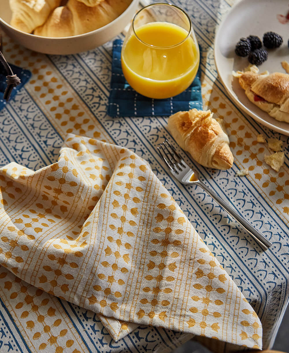 Breakfast scene with a glass of orange juice, croissant, and fruit on a patterned tablecloth