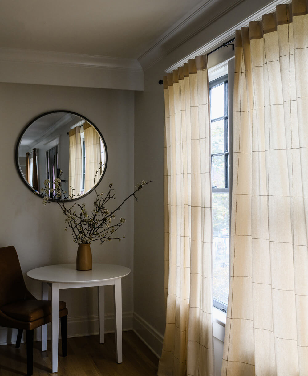 Room interior with a round mirror, small table, and chair near a window with curtains