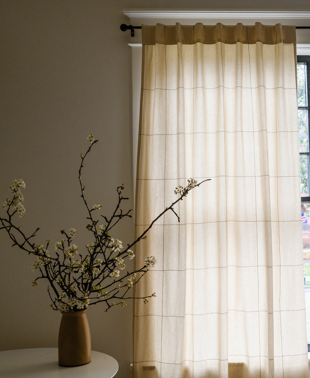 Sheer curtain with a grid pattern in front of a window, with a vase and branches on a table to the left