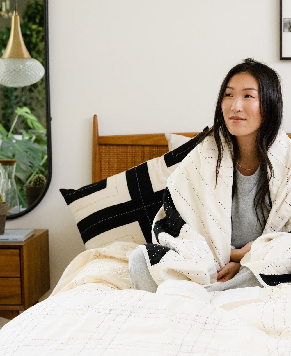 Woman sitting on a bed wrapped in a white blanket with black accents, in a cozy bedroom setting