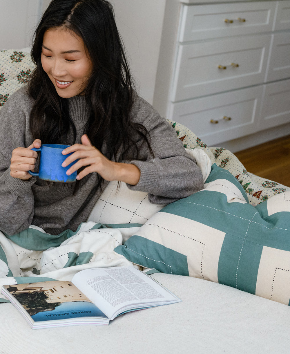 Woman sitting on a couch with a book and mug, surrounded by a cozy blanket and pillows