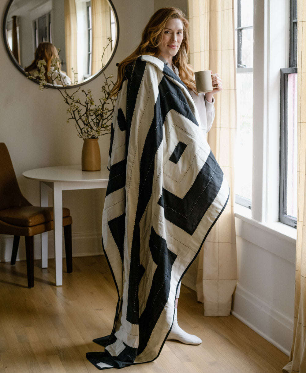 Woman wrapped in a black and white patterned blanket holding a mug in a cozy room.