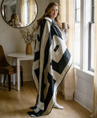 Woman wrapped in a black and white patterned blanket holding a mug in a cozy room.