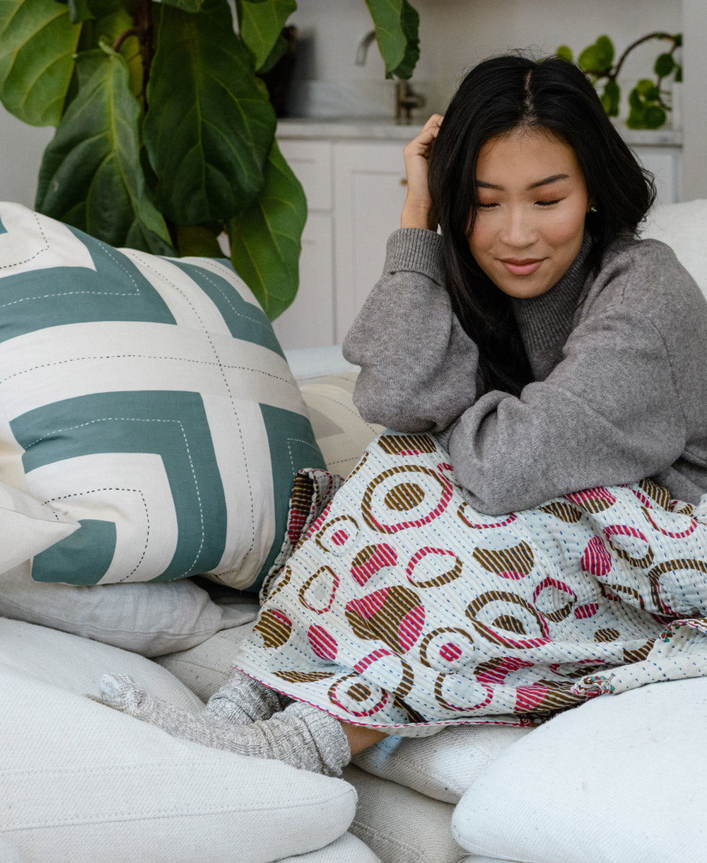 Woman sitting on a couch with a patterned blanket, surrounded by decorative pillows and plants