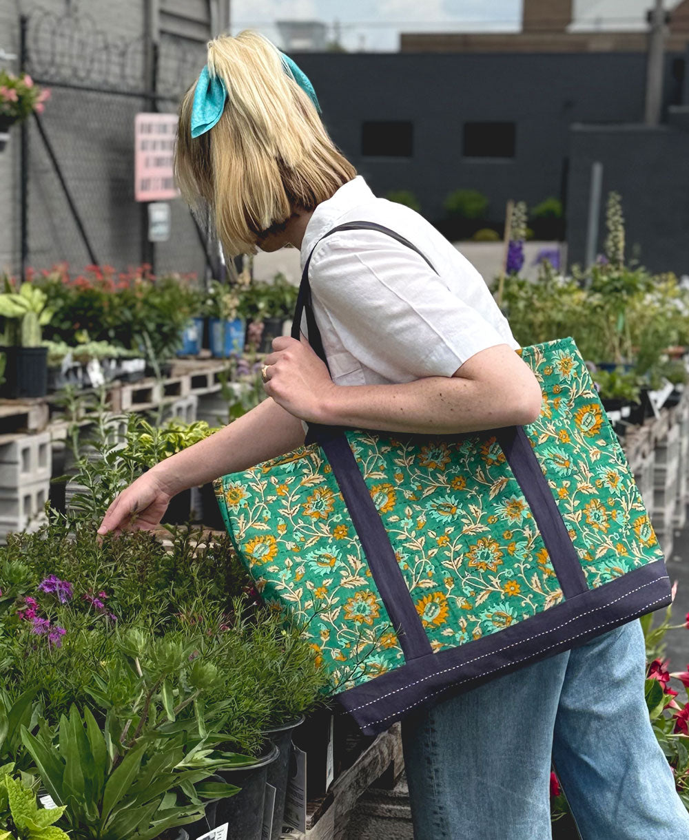 blond woman holding a green floral tote bag in an outdoor setting with plants and buildings in the background