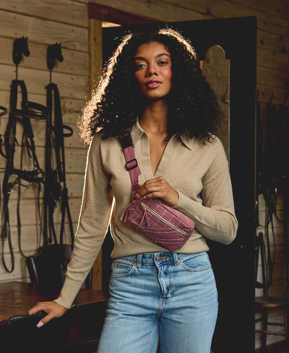 Woman holding a purple crossbody bag in a rustic indoor setting with wooden walls and horse equipment
