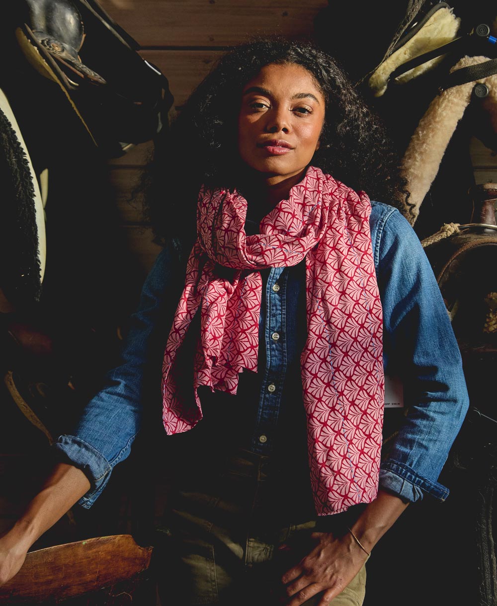 woman wearing a pink patterned long cotton scarf and denim button up shirt in a rustic setting with horses