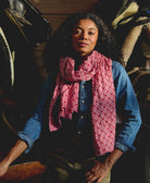 woman wearing a pink patterned long cotton scarf and denim button up shirt in a rustic setting with horses