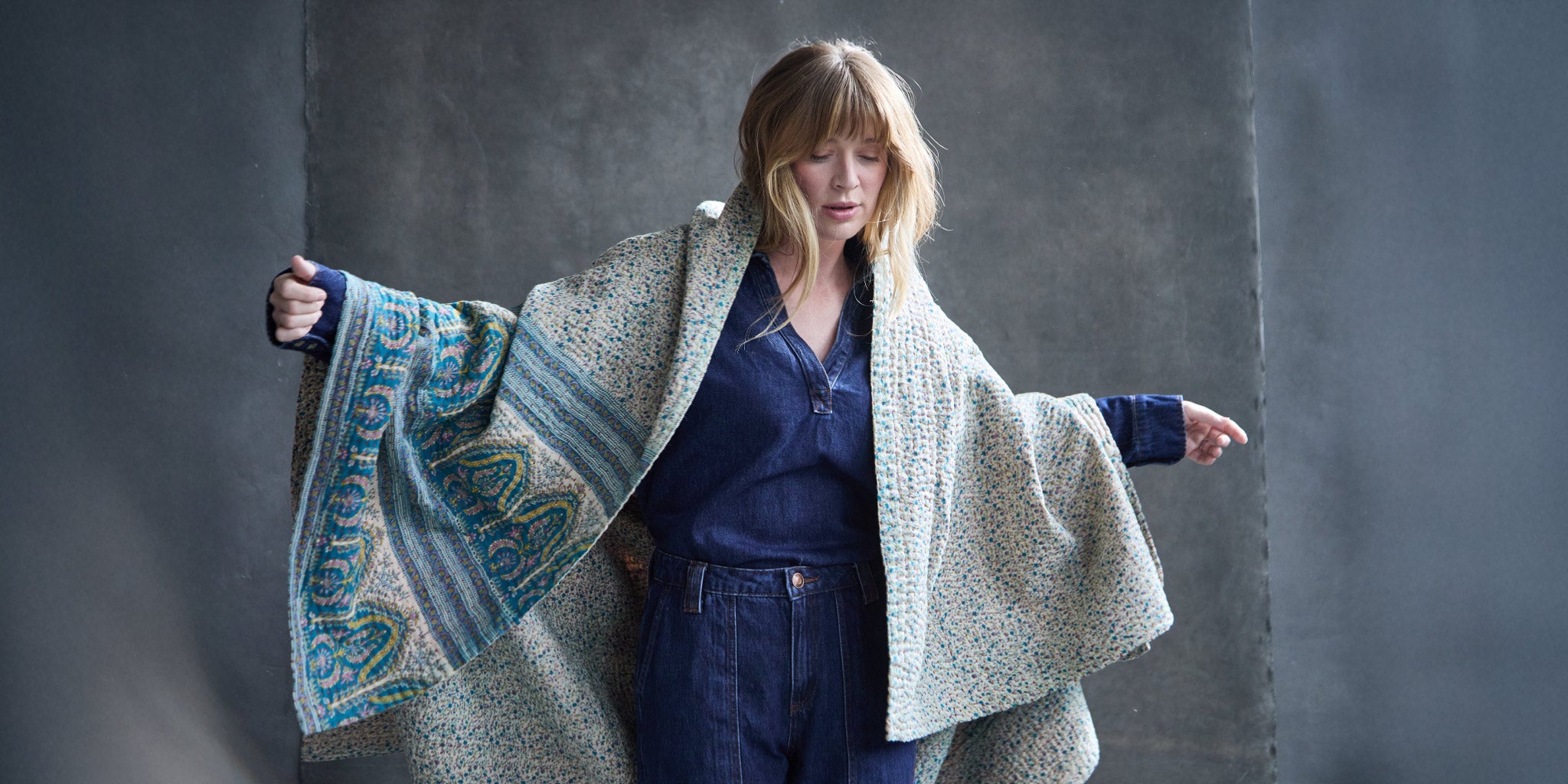 Woman wearing a patterned kantha quilt over a plain garment against a gray background