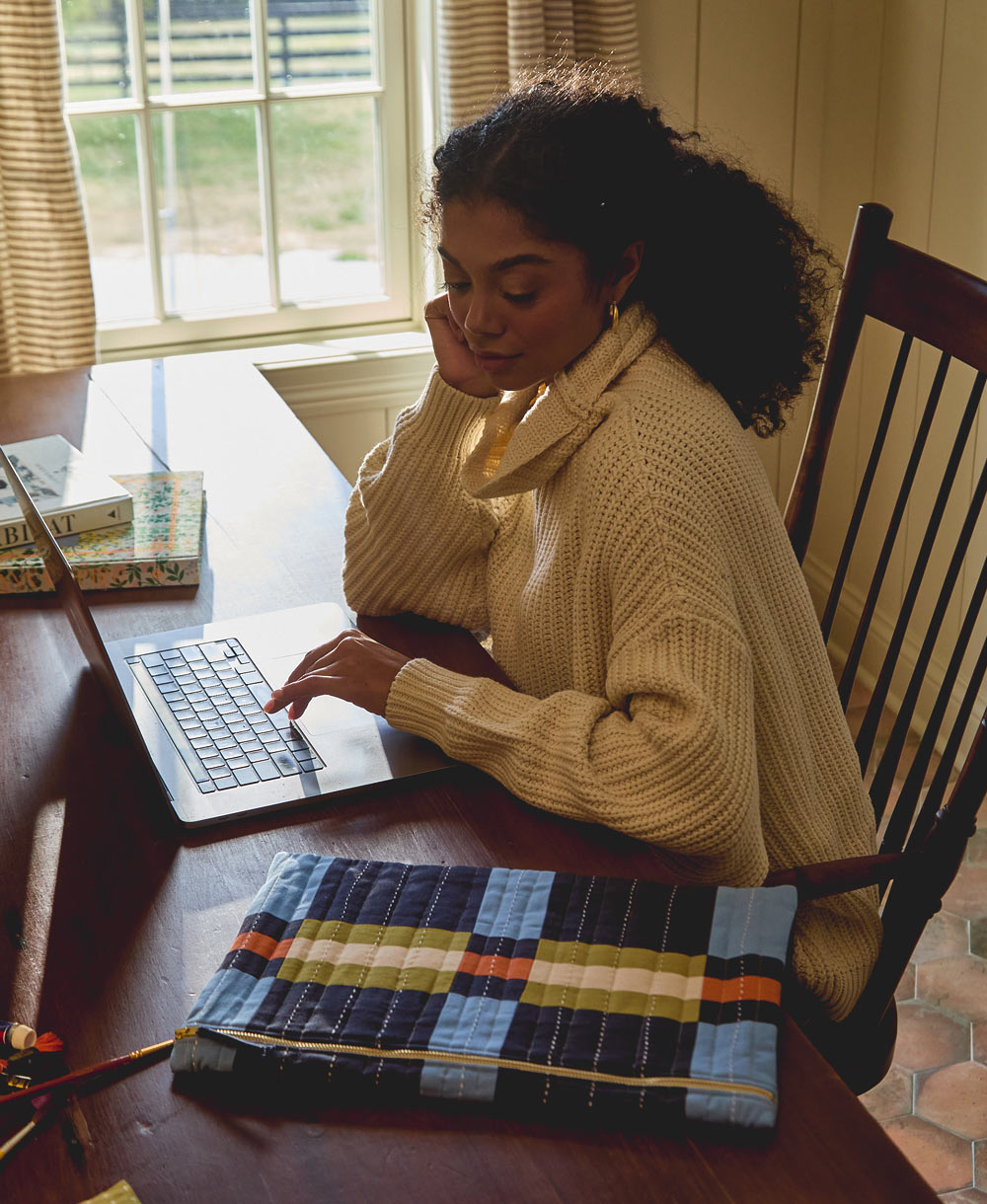 woman sitting at wooden desk working on laptop with a quilted plaid laptop sleeve next to the computer
