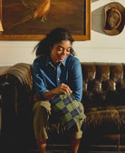 Woman sitting on a brown leather couch holding a plaid bag, with a painting of a bird and a cowboy hat in the background