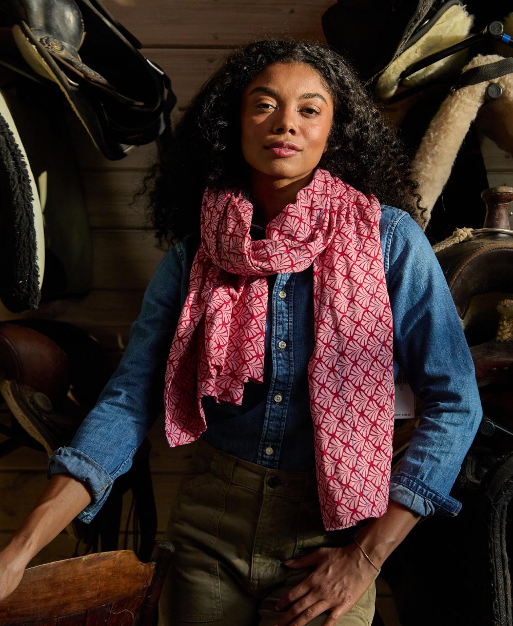 woman wearing a pink patterned scarf and blue jacket in an equestrian setting with saddles in the background