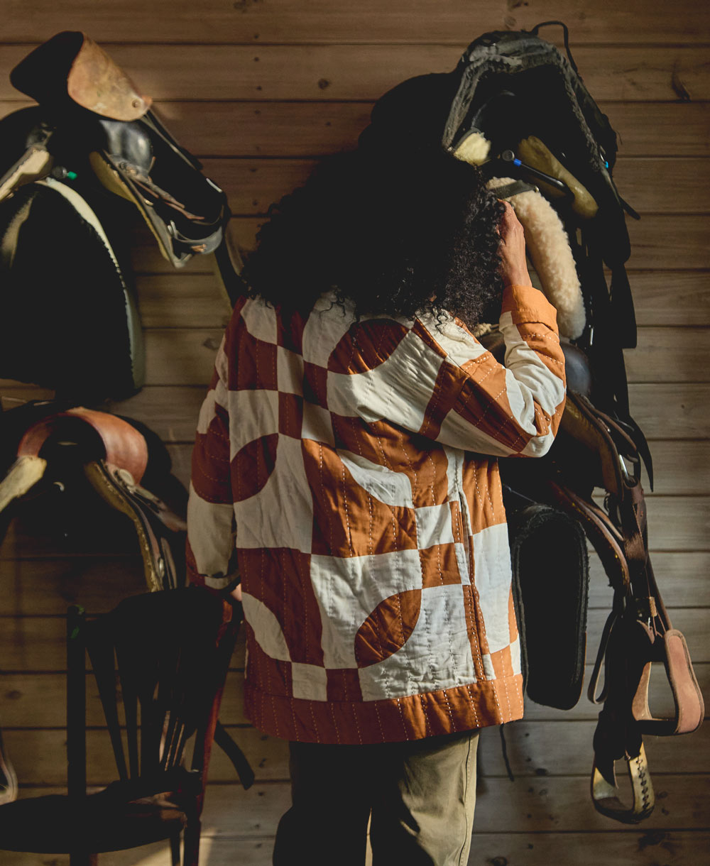 woman wearing a checkered quilted jacket standing in a room with horse saddles on the wall