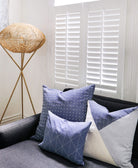 Decorative pillows on a sofa with a woven floor lamp and white shutters in the background