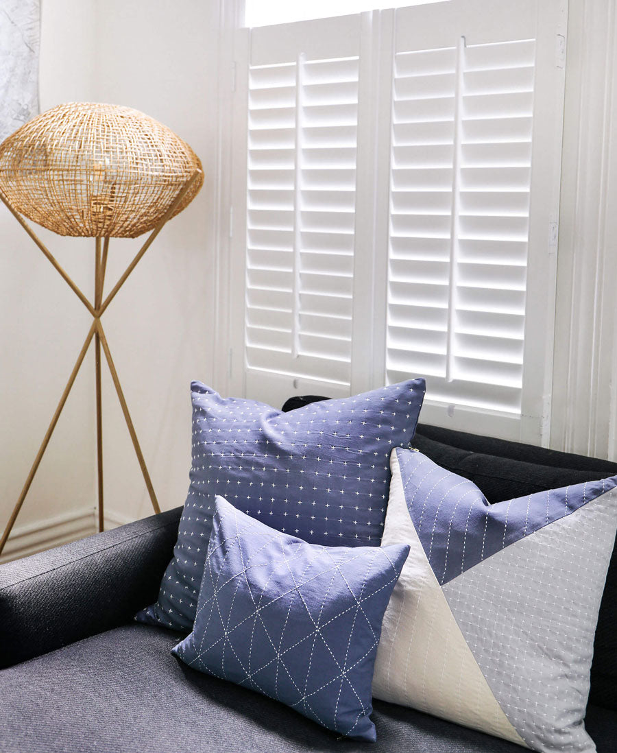 Decorative pillows on a sofa with a woven floor lamp and white shutters in the background