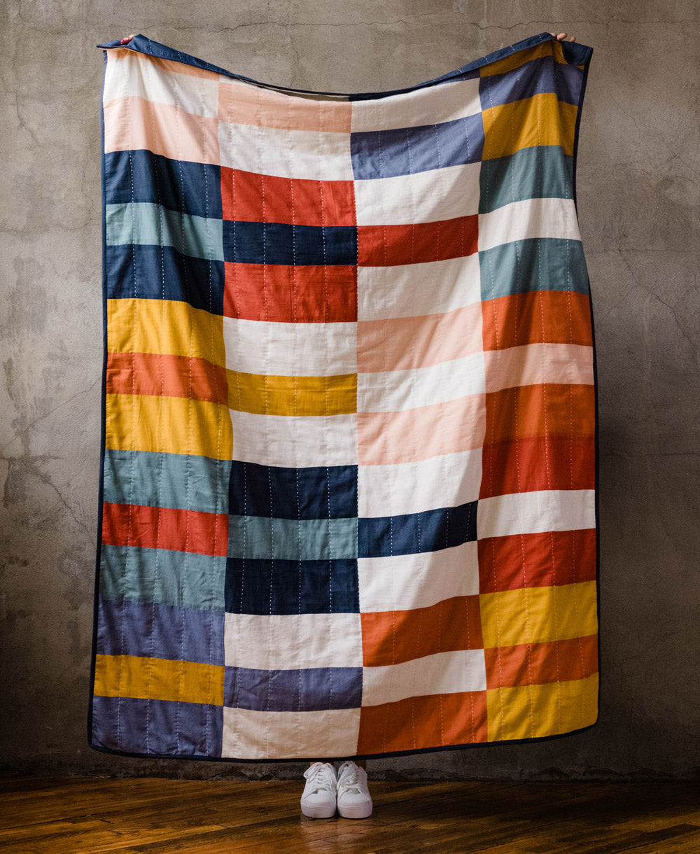 A colorful checkered quilt draped over a person against a concrete wall.