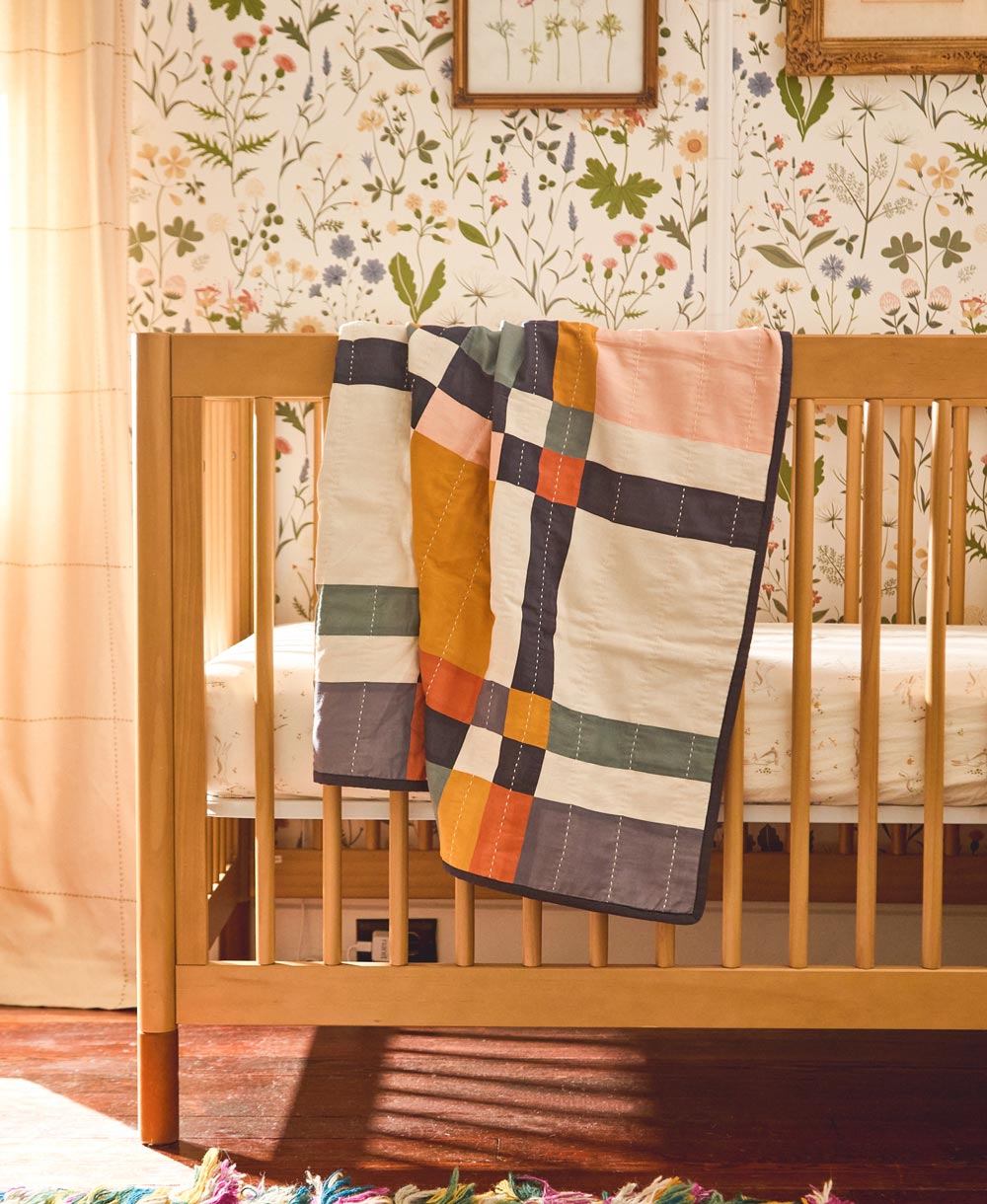 Patchwork quilt draped over a wooden crib with floral wallpaper in the background