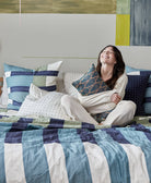 Woman sitting on a bed with colorful bedding and pillows in a room with abstract art on the wall