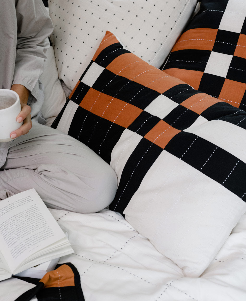 Person sitting on a bed with checkered pillows and a book, holding a white mug