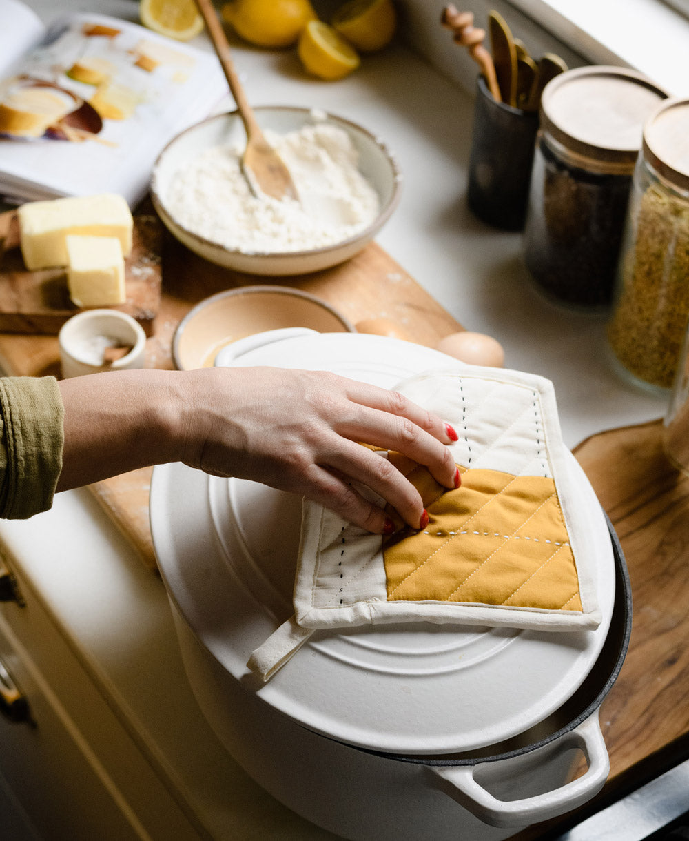 Person holding a yellow oven mitt over a white pot on a kitchen counter with various kitchen items