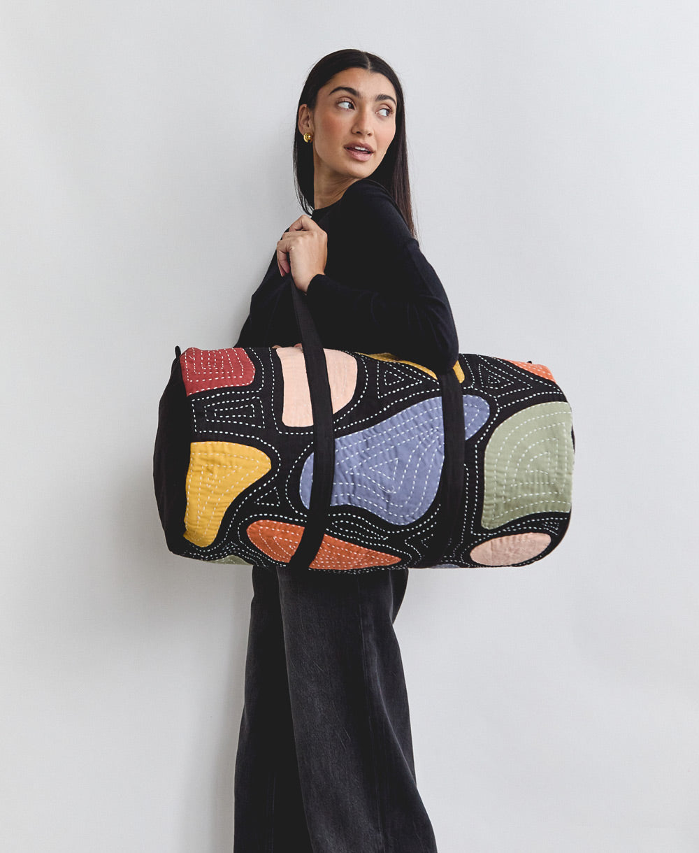 woman carrying black weekender duffle bag over shoulder with bold colorful patchwork applique