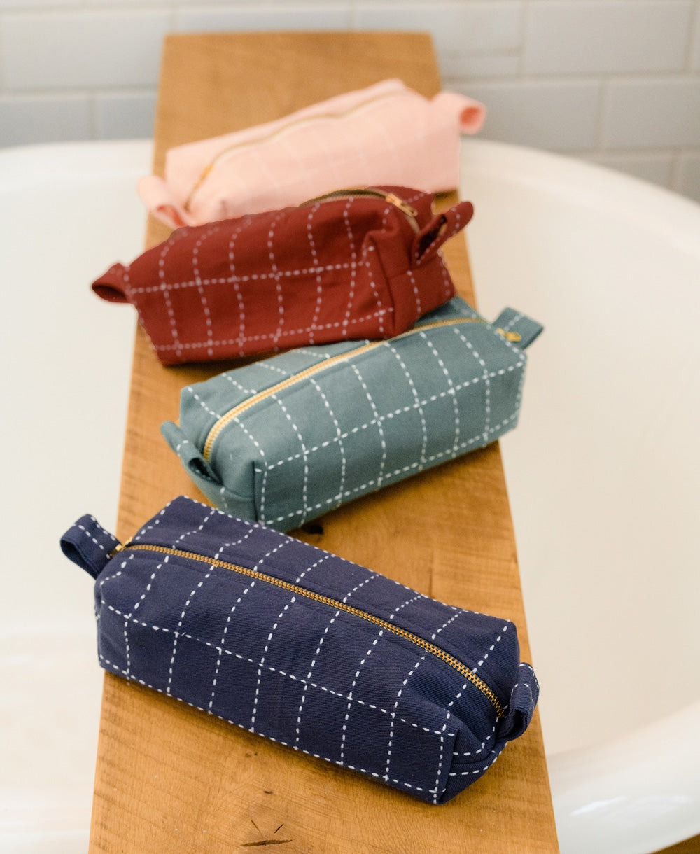 grouping of 4 small toiletry bags in navy, spruce, rust and pink sitting on wooden bath table