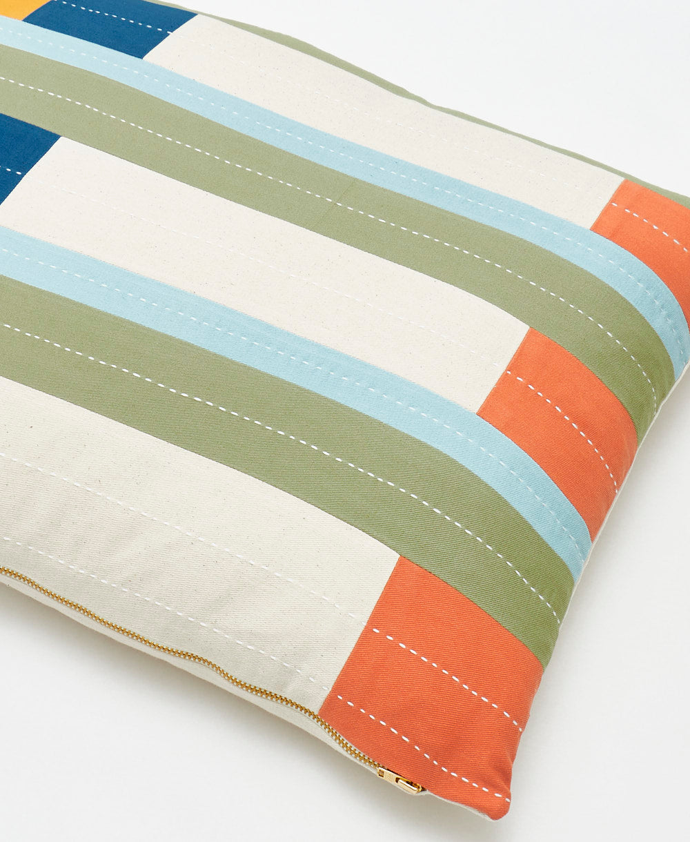 Colorful striped pillow in shades of orange, sky blue, sage green, and cobalt blue with a geometric pattern on a white background