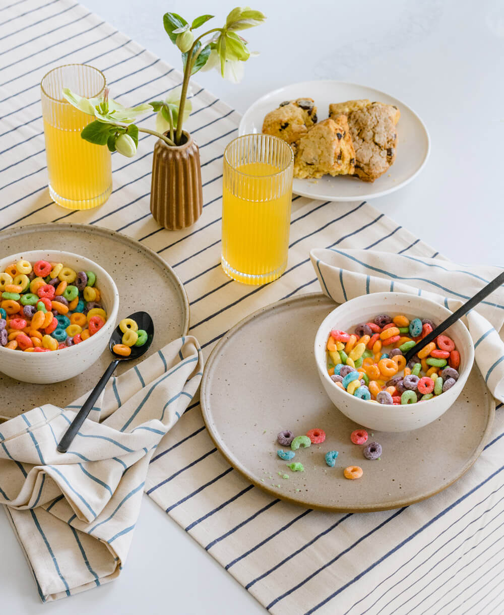 Two bowls of colorful cereal with a glass of orange juice on a striped tablecloth