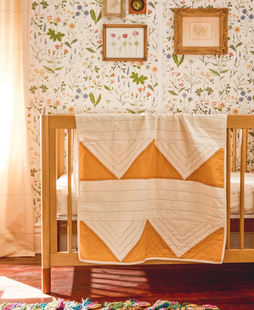 Crib with a quilt draped over it against a floral wallpapered wall.