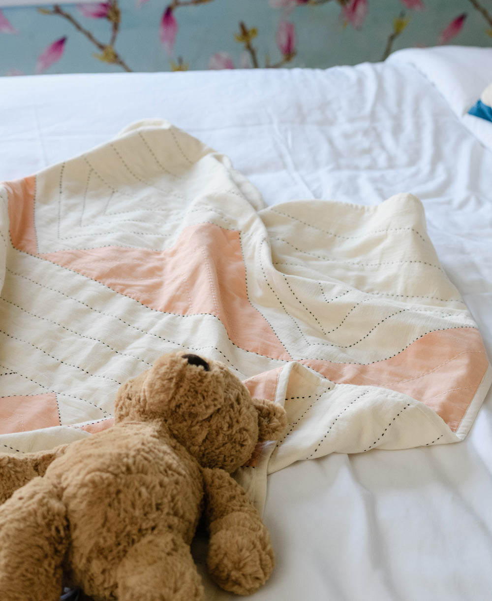 Brown teddy bear under a patterned quilt on a bed with floral wallpaper.