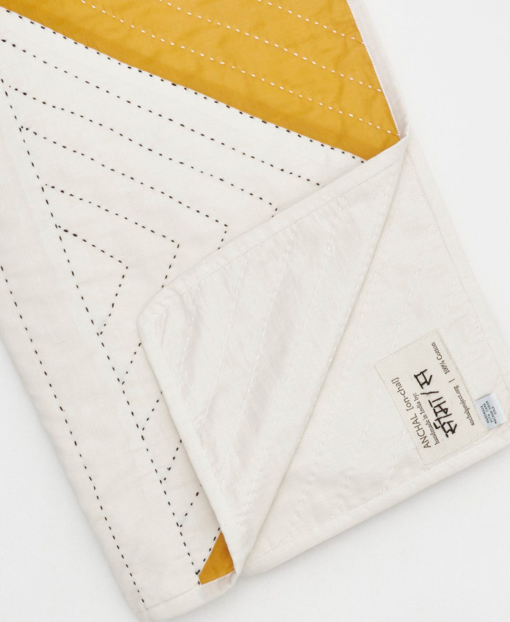 Close-up of a quilt with white and mustard yellow fabric, featuring Anchal's artisan signed tag